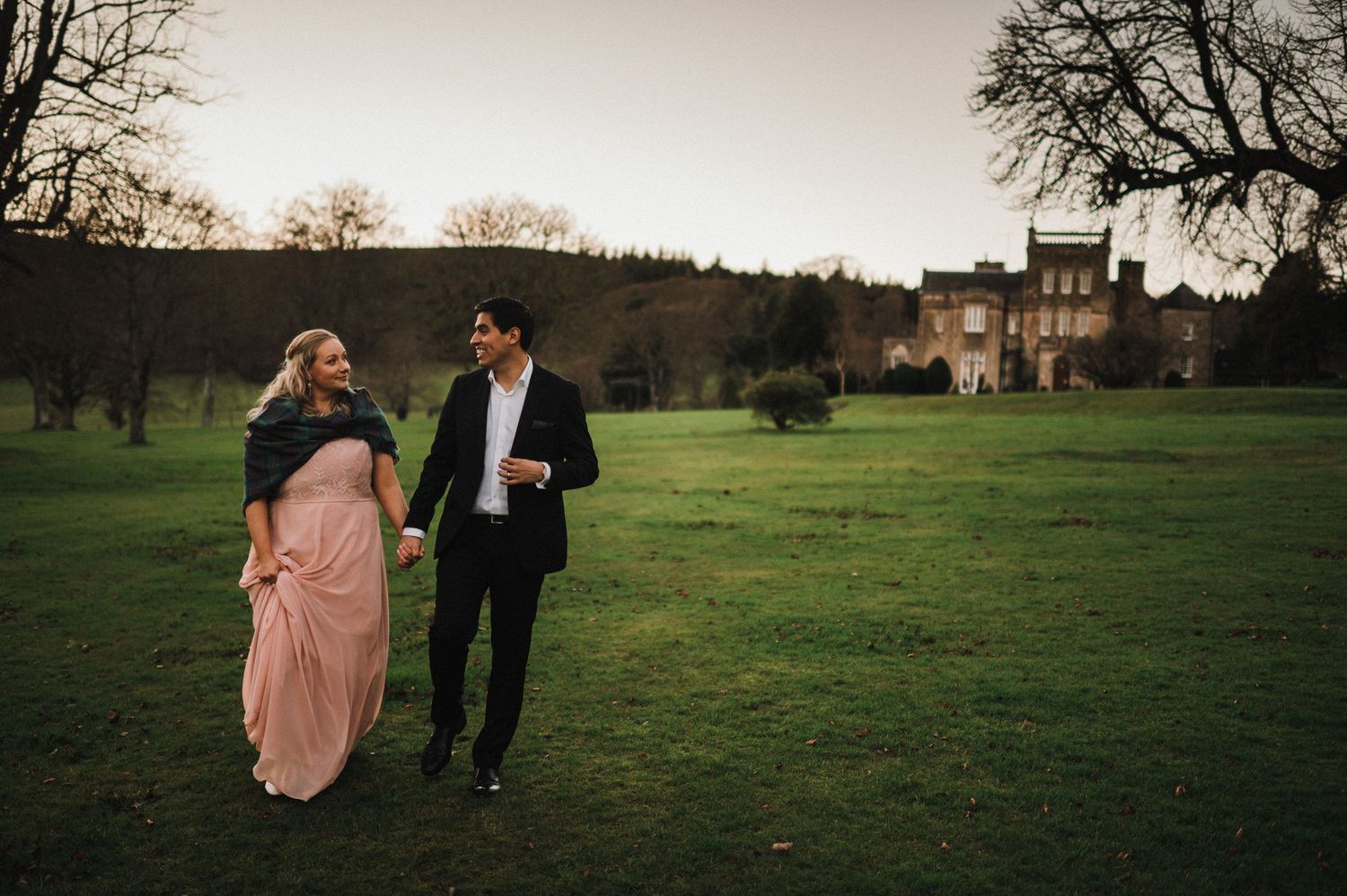 A couple walks together on grass with a historic mansion visible in the background.