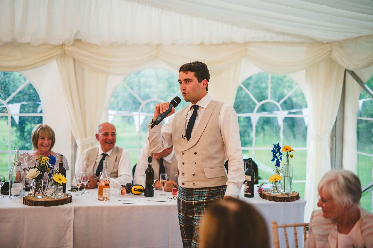 Wedding speaker in traditional Scottish attire giving speech at reception dinner.