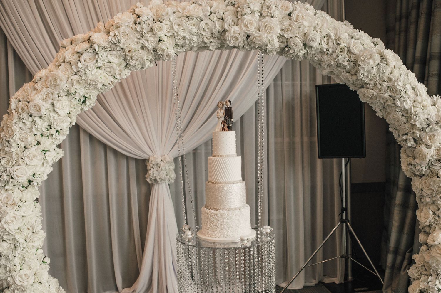 White floral arch with draped fabric frames an elegant wedding cake on a crystal stand.