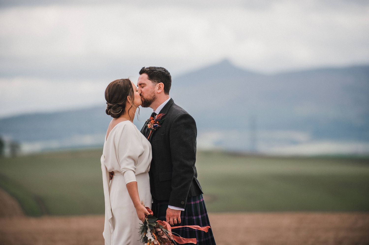A romantic wedding moment captured with mountains in the background and traditional Scottish dress.