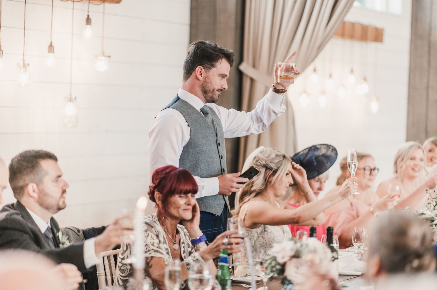 A wedding speech moment captured with guests cheering and raising hands in a venue with pendant lighting.