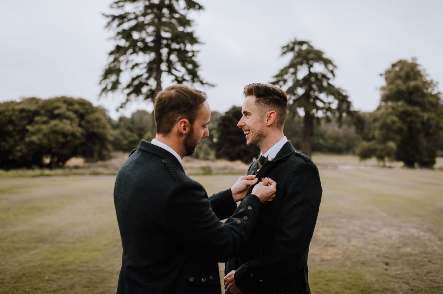 Two men in formal suits adjusting boutonnieres on a grassy lawn.