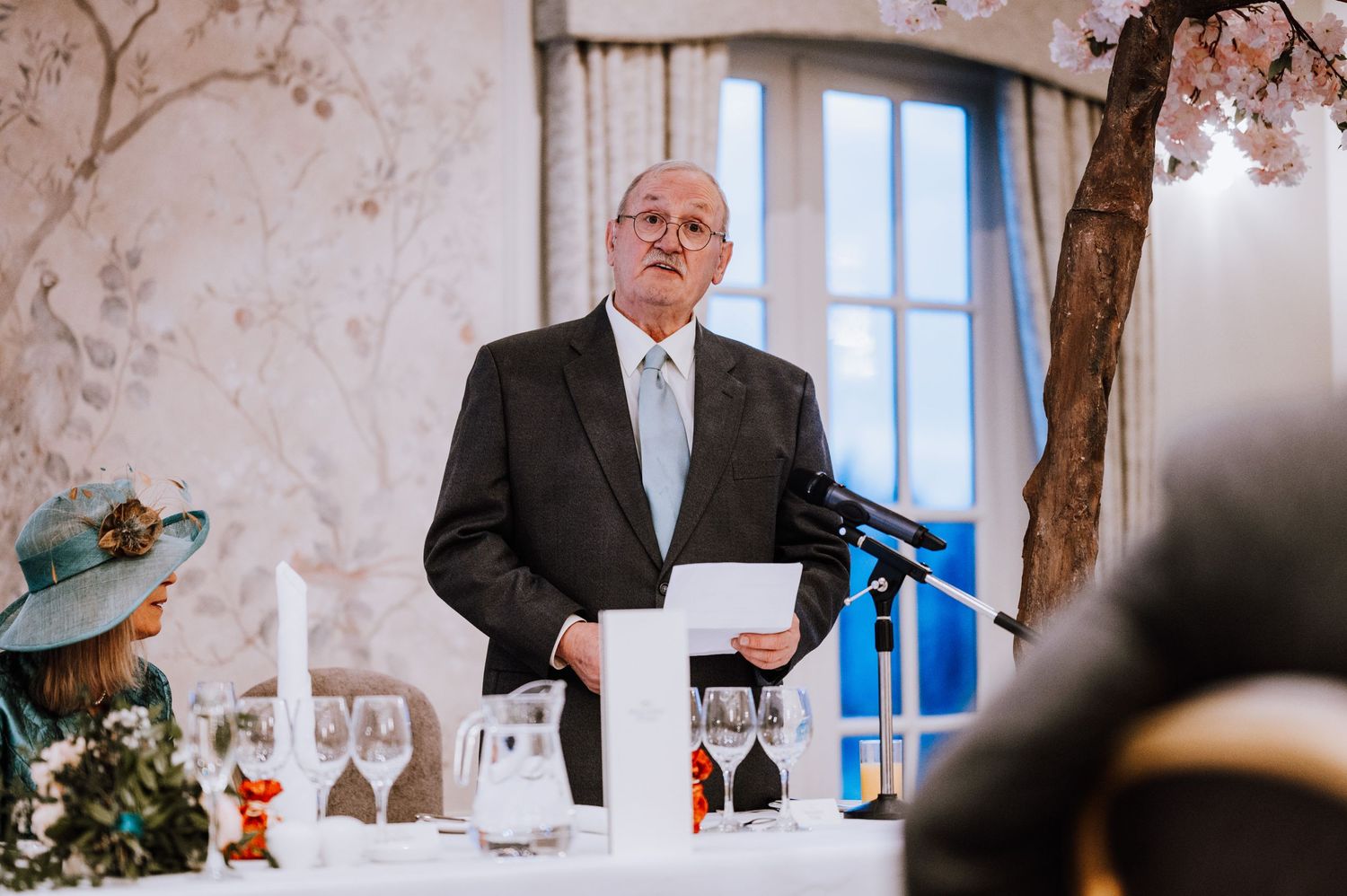 Speaker giving a toast at formal reception with decorative branches and white tablecloths.