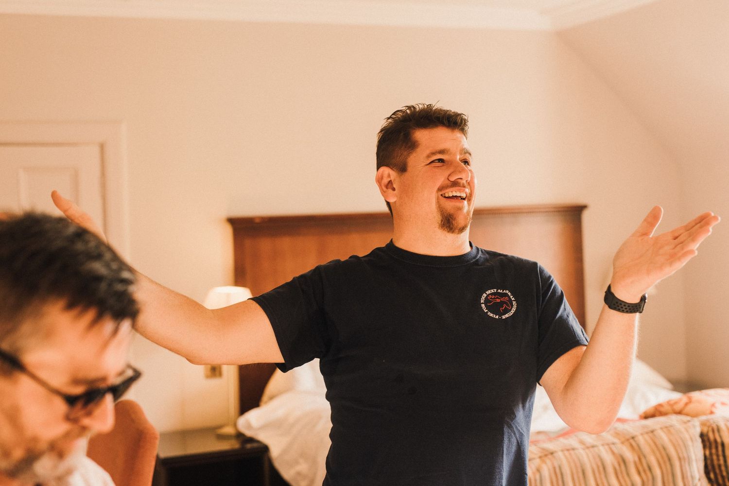 Person in black t-shirt gesturing with arms spread wide in an energetic pose indoors.