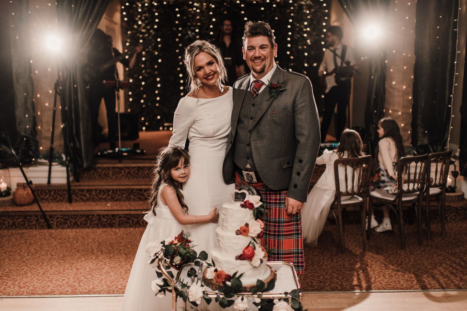A Scottish wedding celebration with the couple posing in front of twinkling lights, featuring a traditional kilt and white dress.