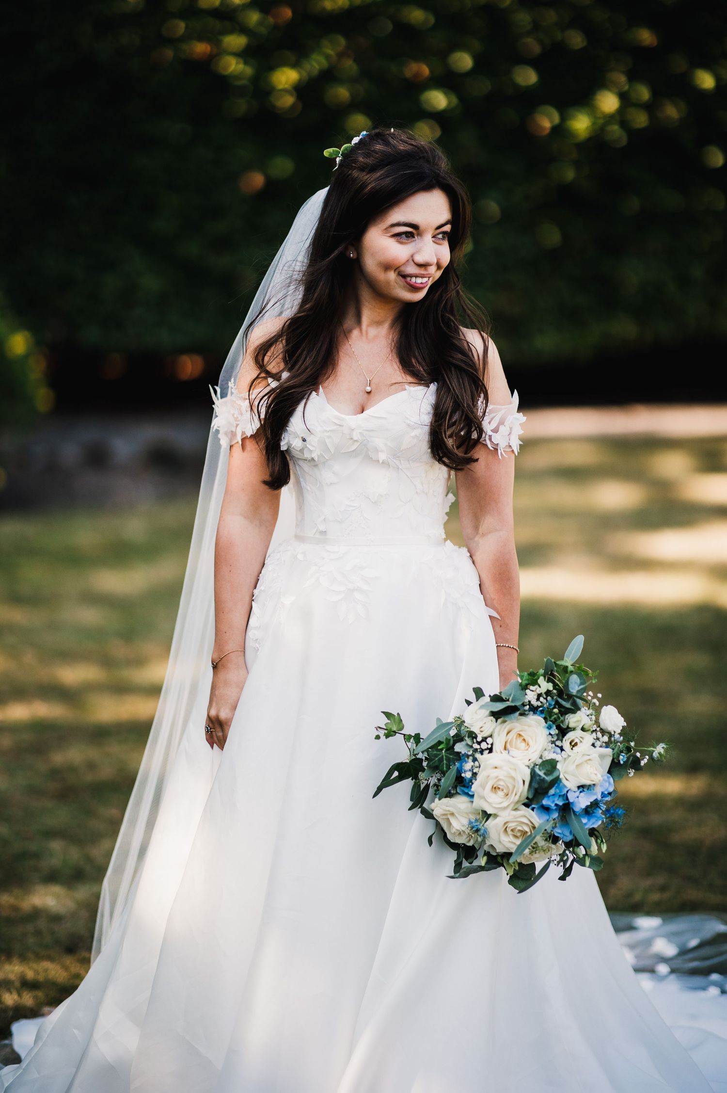 Bride in flowing white gown holds elegant blue and white floral bouquet in garden setting.
