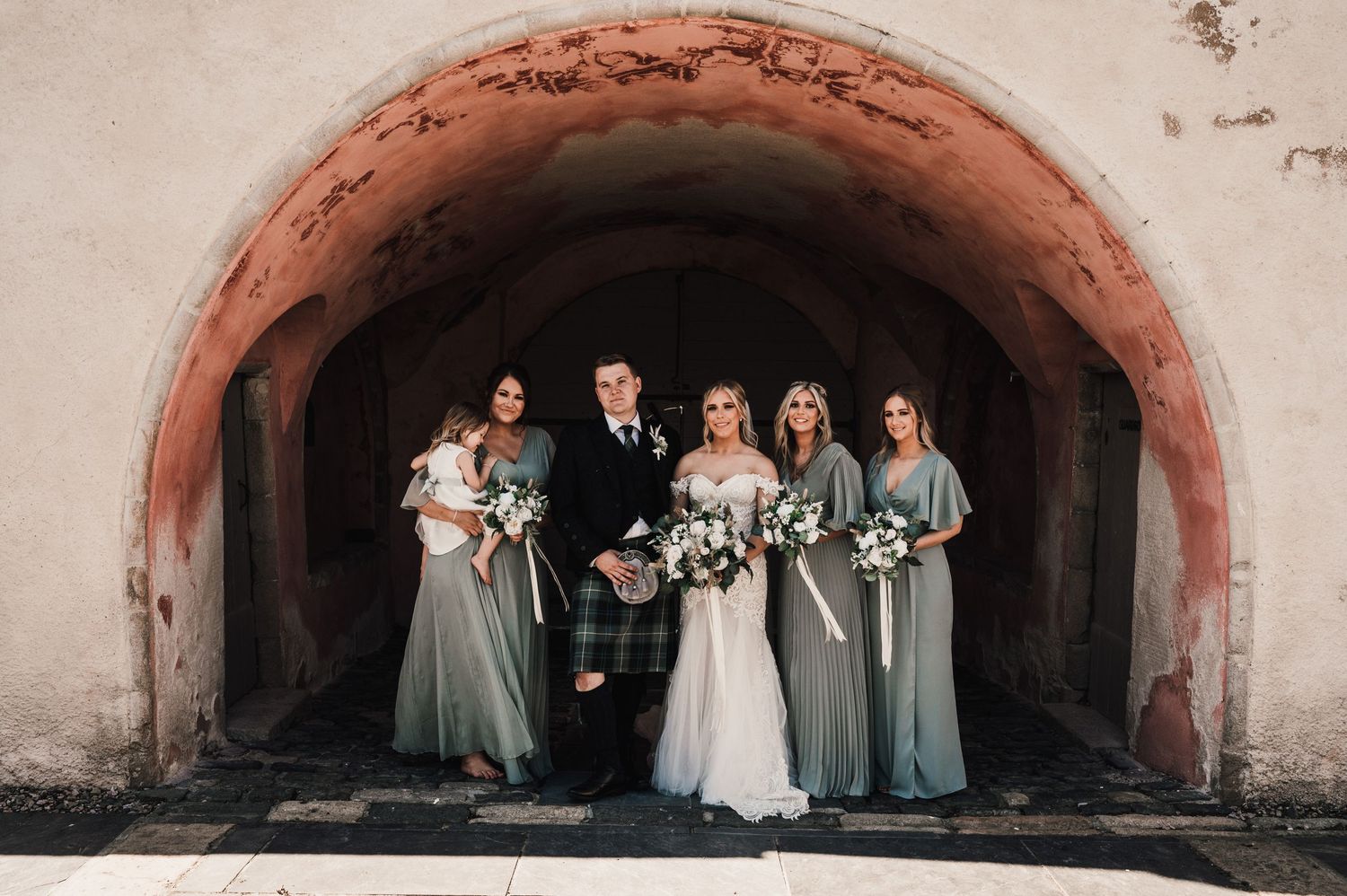 Intimate wedding party photo under rustic stone archway with sage green bridesmaid dresses.