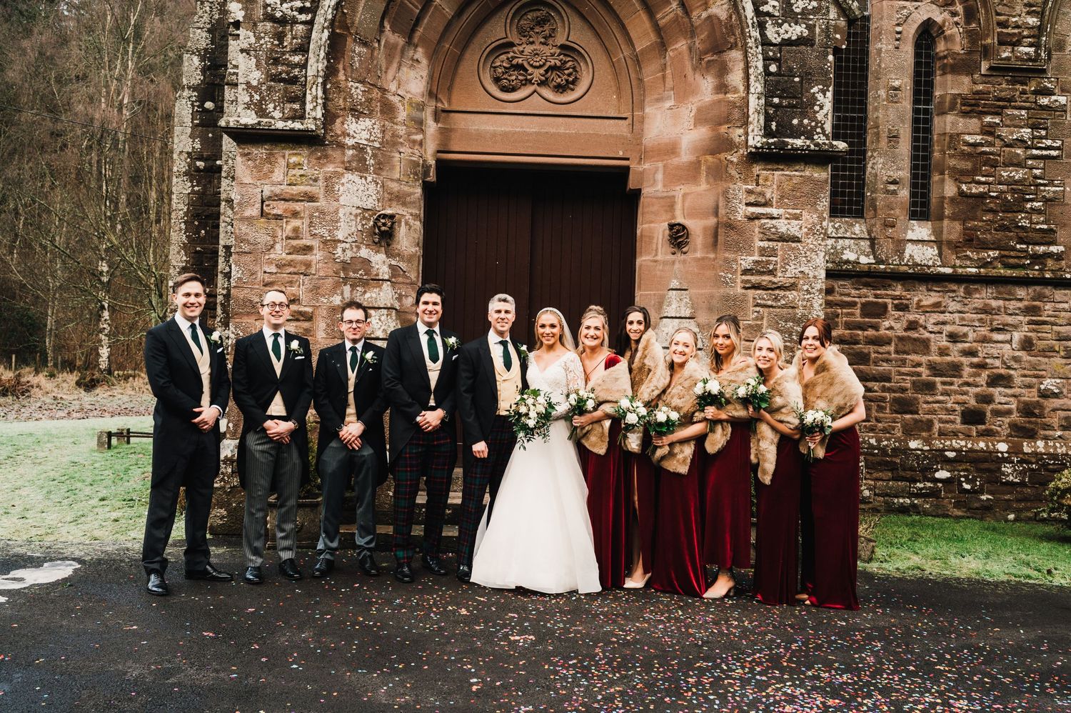 Wedding party in burgundy and black formal attire lined up at stone church entrance.