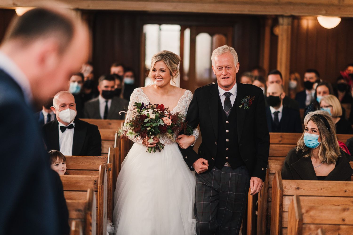 A wedding processional in a traditional church with wooden pews and masked attendees.