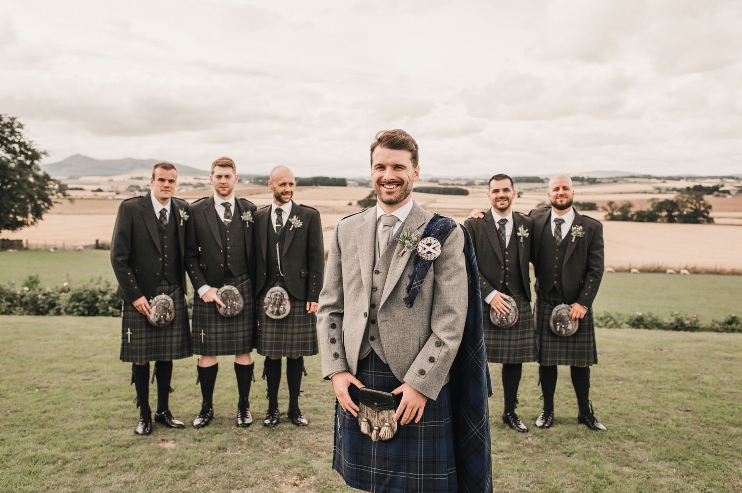 Wedding party in kilts with groom in light suit standing on scenic hilltop.