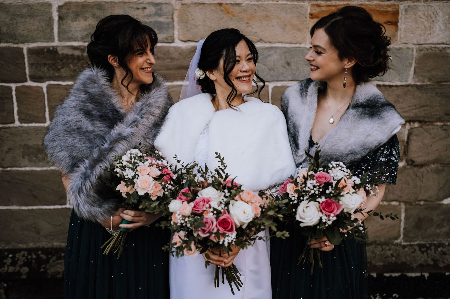 Three wedding attendants in grey shawls hold pink and white floral bouquets against stone wall.