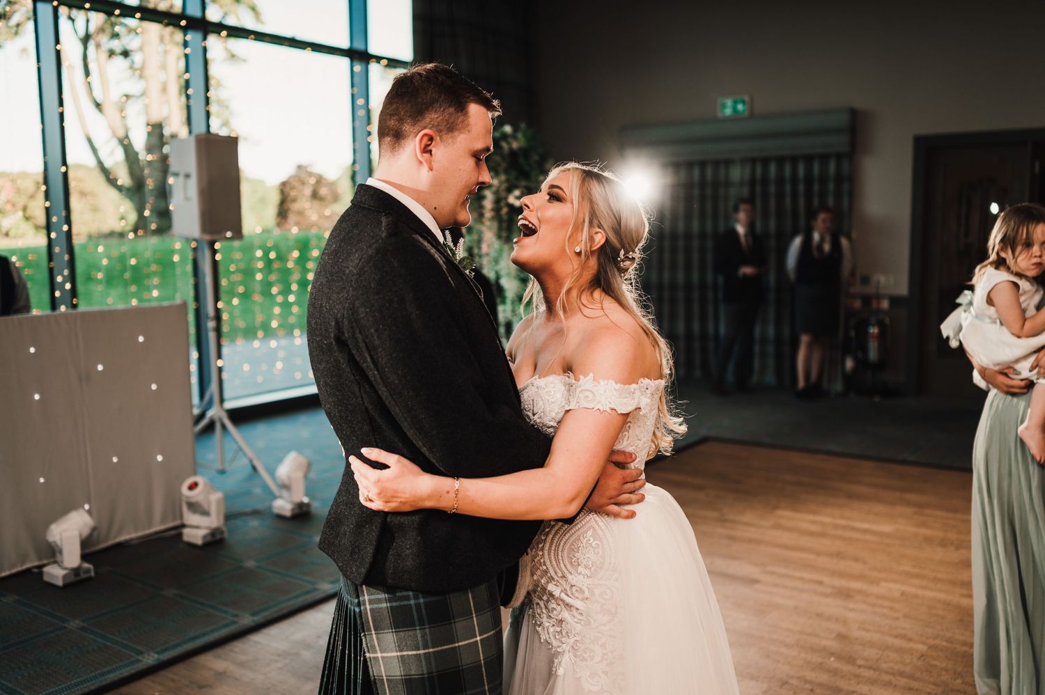 Romantic first dance moment in a reception hall with twinkling lights and large windows.