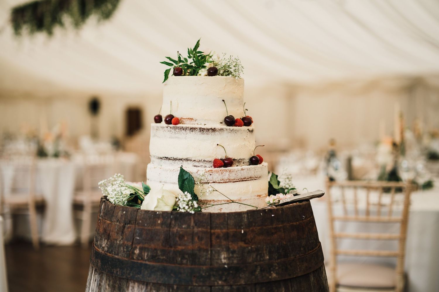 Rustic three-tier white wedding cake displayed on wooden barrel with fresh berries and greenery.