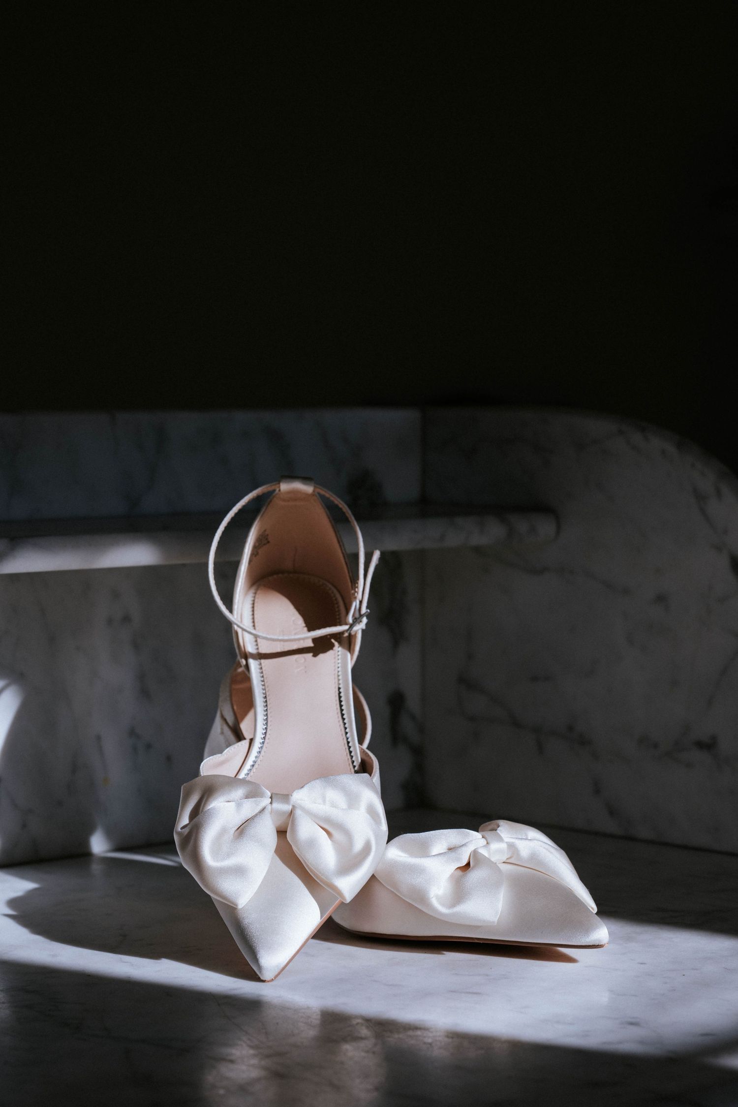 White satin bridal shoes with bows displayed on a marble surface in dramatic lighting.