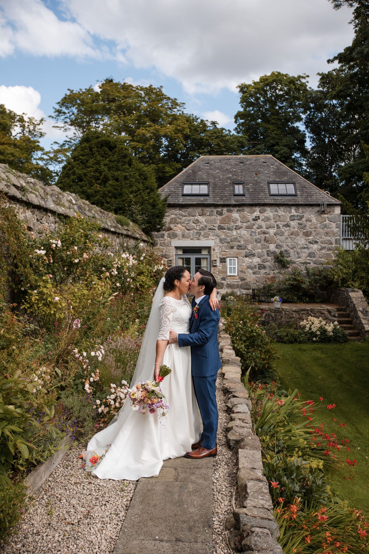 A couple shares a romantic moment in front of a rustic stone cottage with a garden path and flowering plants.
