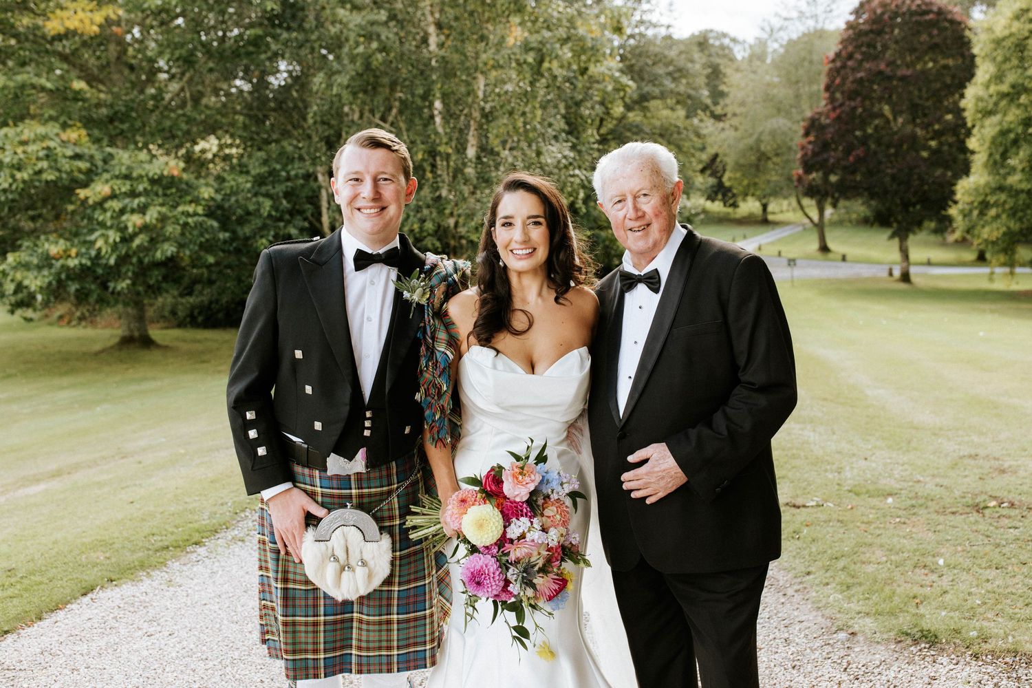 Elegant wedding portrait featuring traditional Scottish formal wear in lush garden setting.
