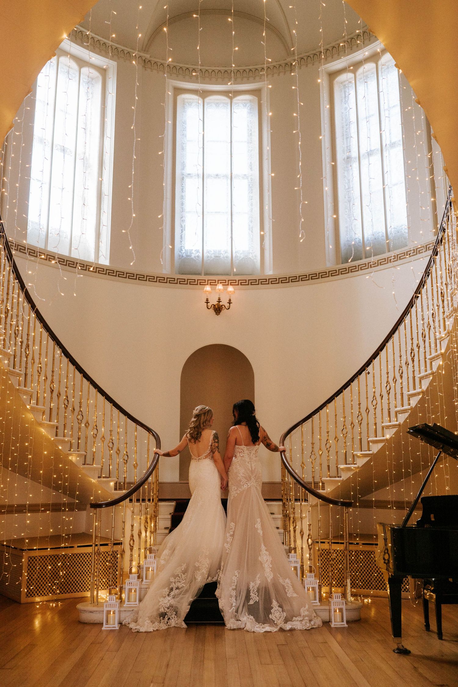 Two wedding dresses on display in an elegant staircase hall with twinkling fairy lights and a grand piano.