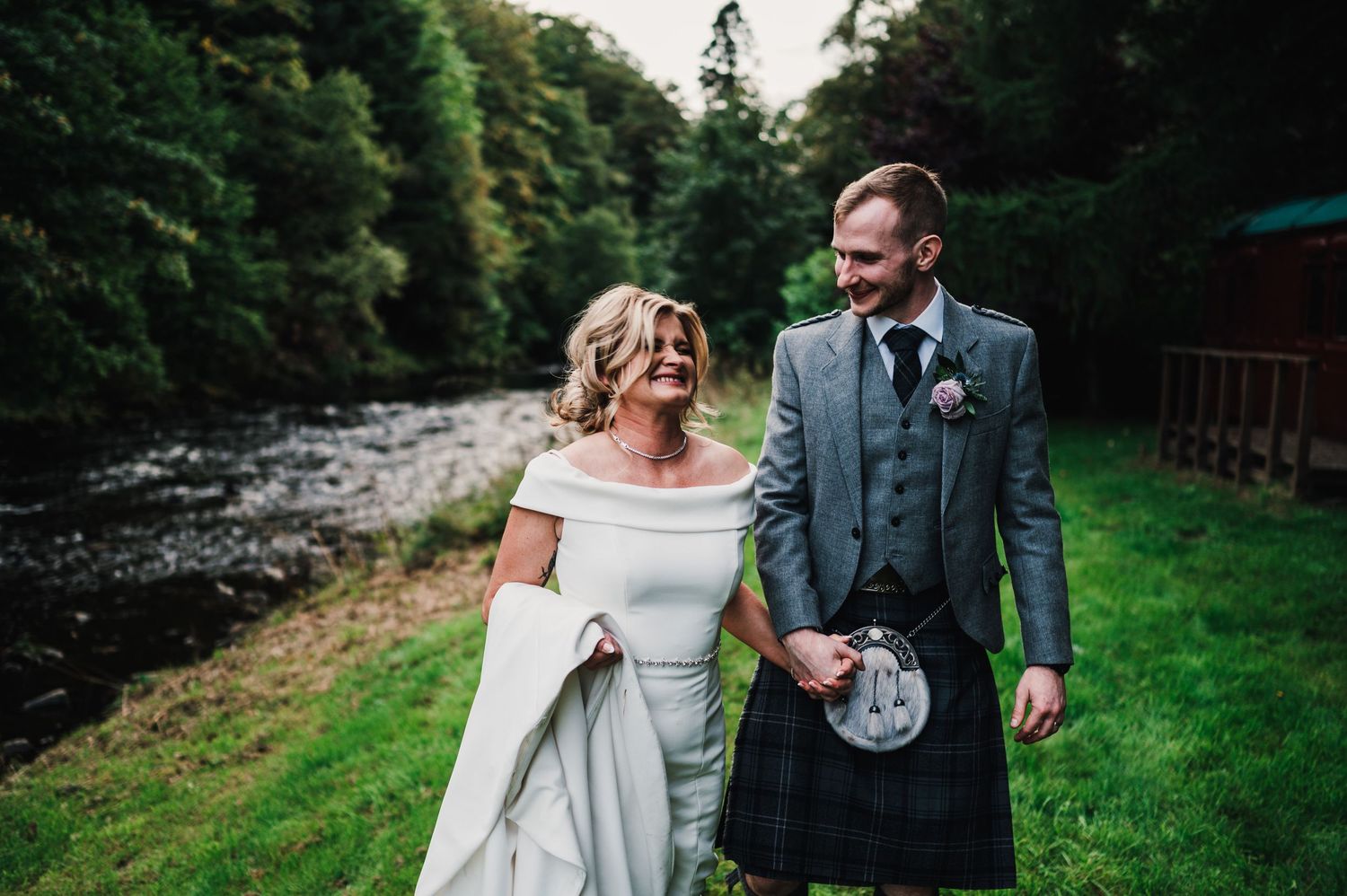Newlyweds walk hand in hand through a wooded path surrounded by greenery.