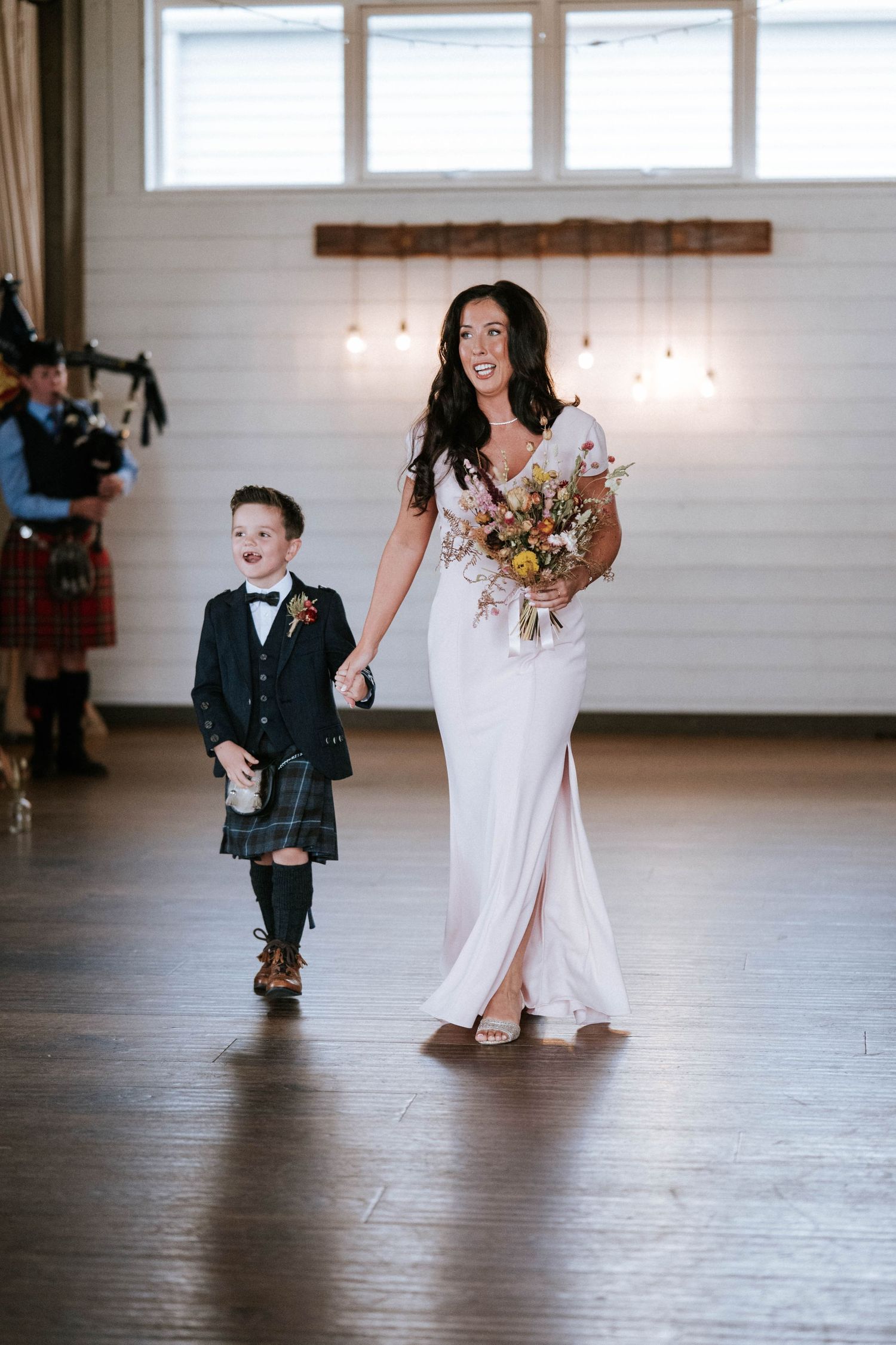 A young ring bearer in a kilt walks down the aisle with the bride holding a bouquet.