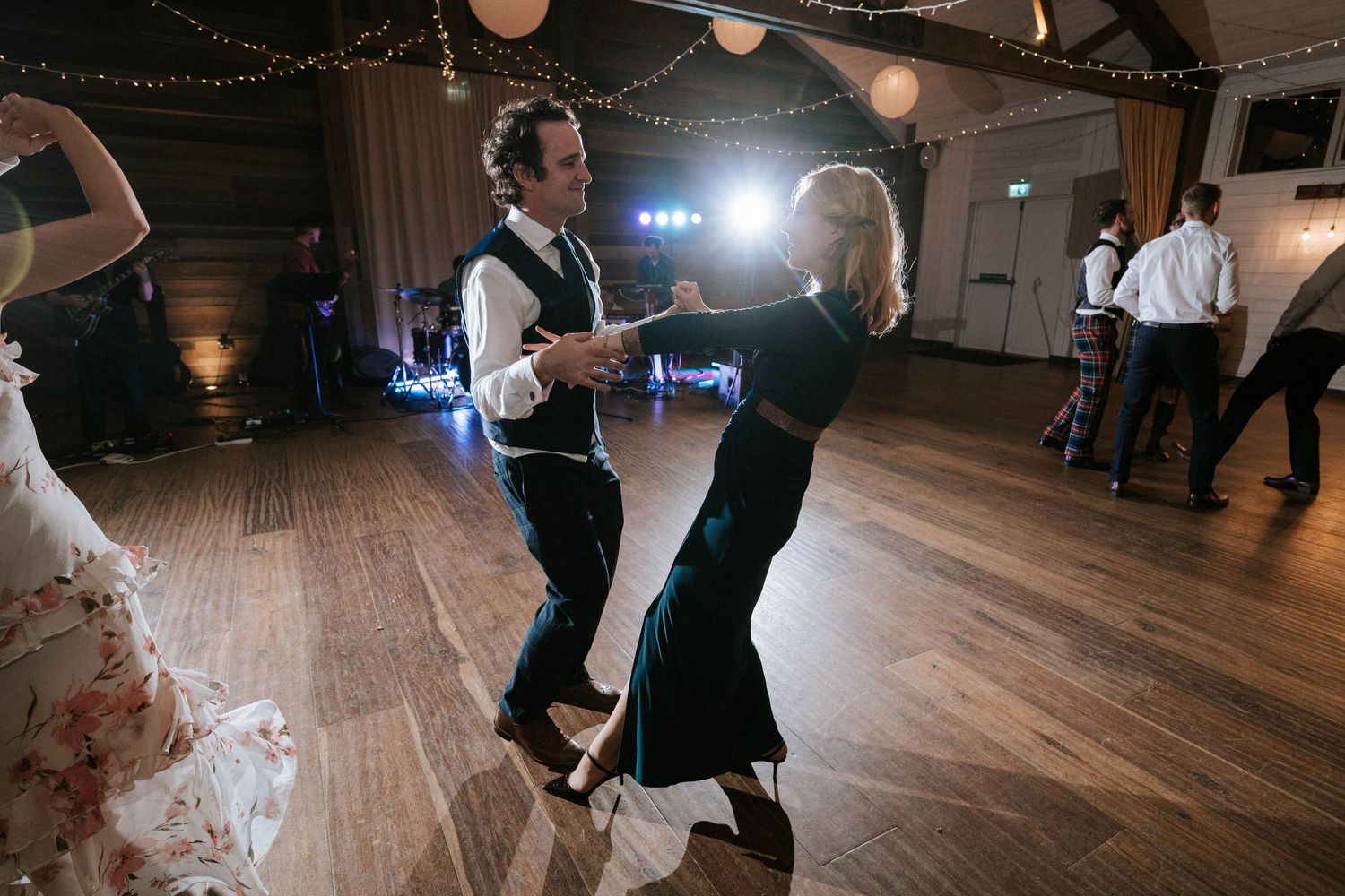 Couples dancing on wooden floor under string lights at wedding reception.