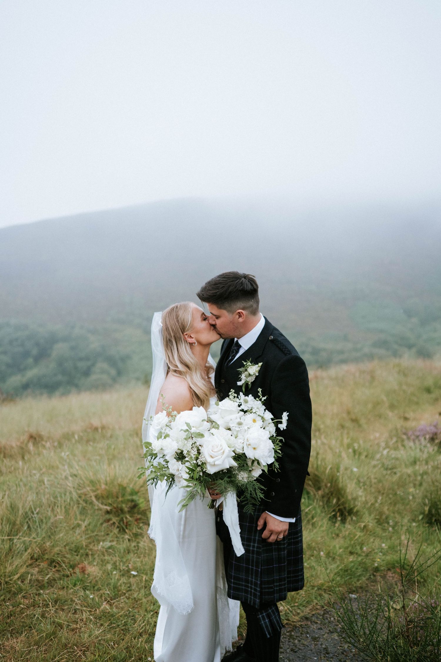 Romantic wedding couple embraces on misty Scottish hillside with white bouquet.