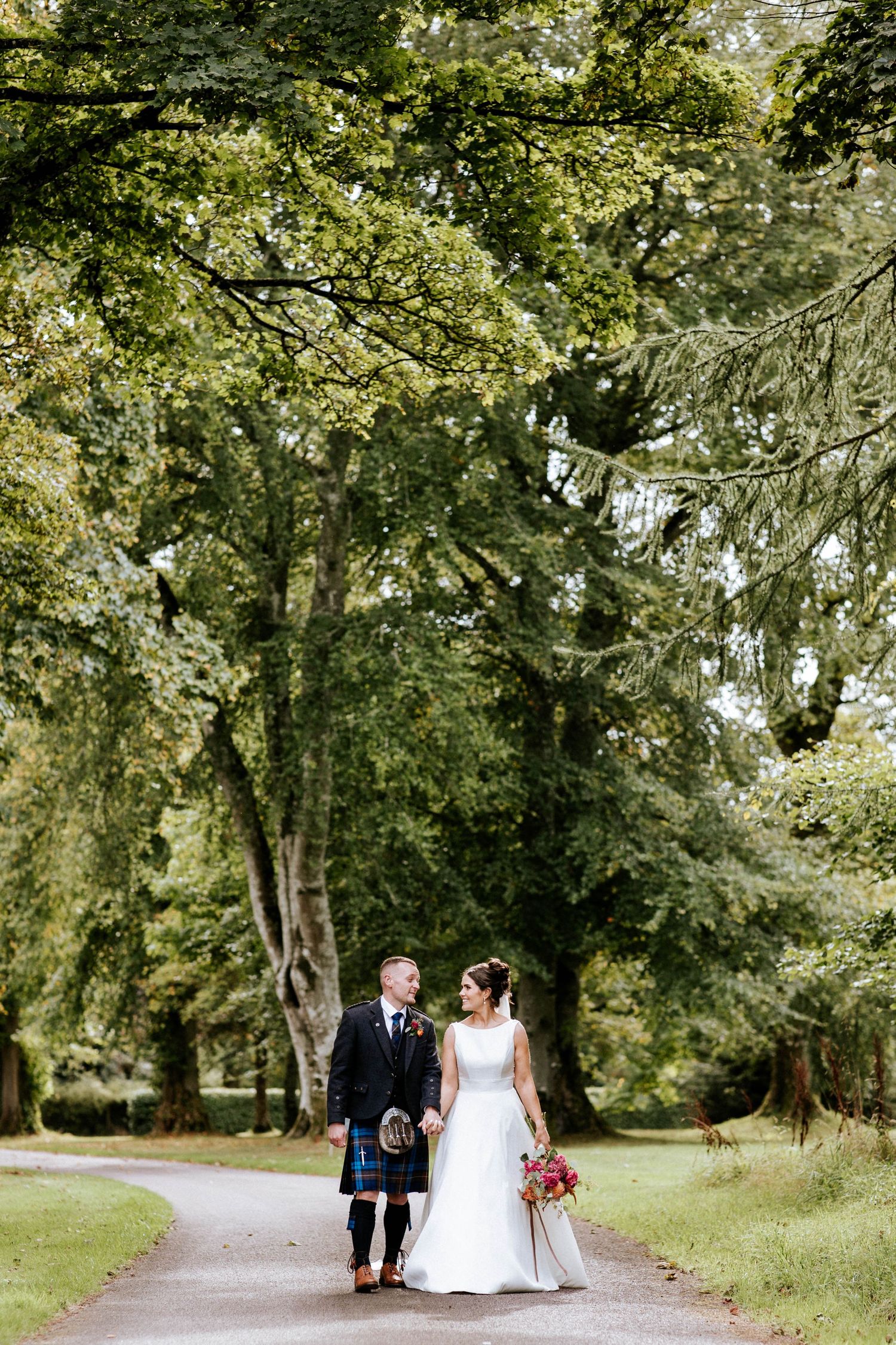 A couple walks down a tree-lined path in a lush garden setting during their wedding.