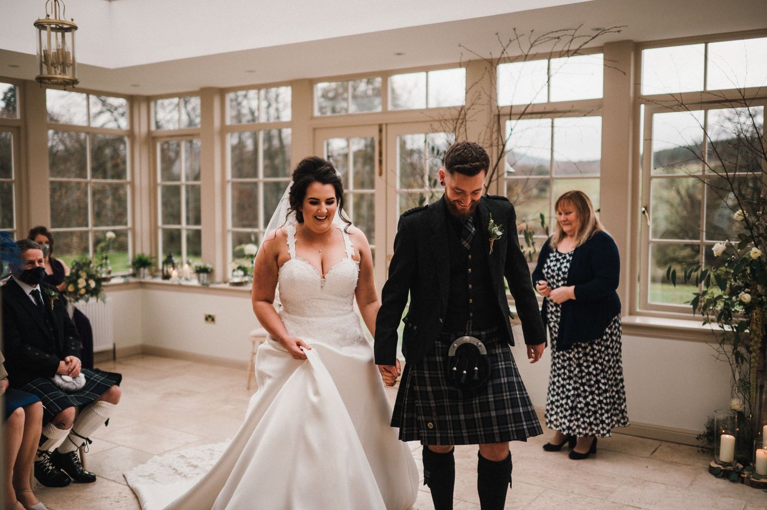 Couple in wedding dress and kilt walk through bright conservatory during Scottish ceremony.
