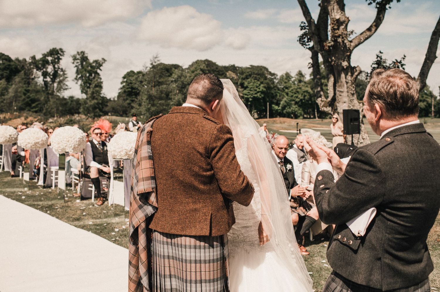 An emotional outdoor wedding ceremony moment with people wearing traditional Scottish attire.