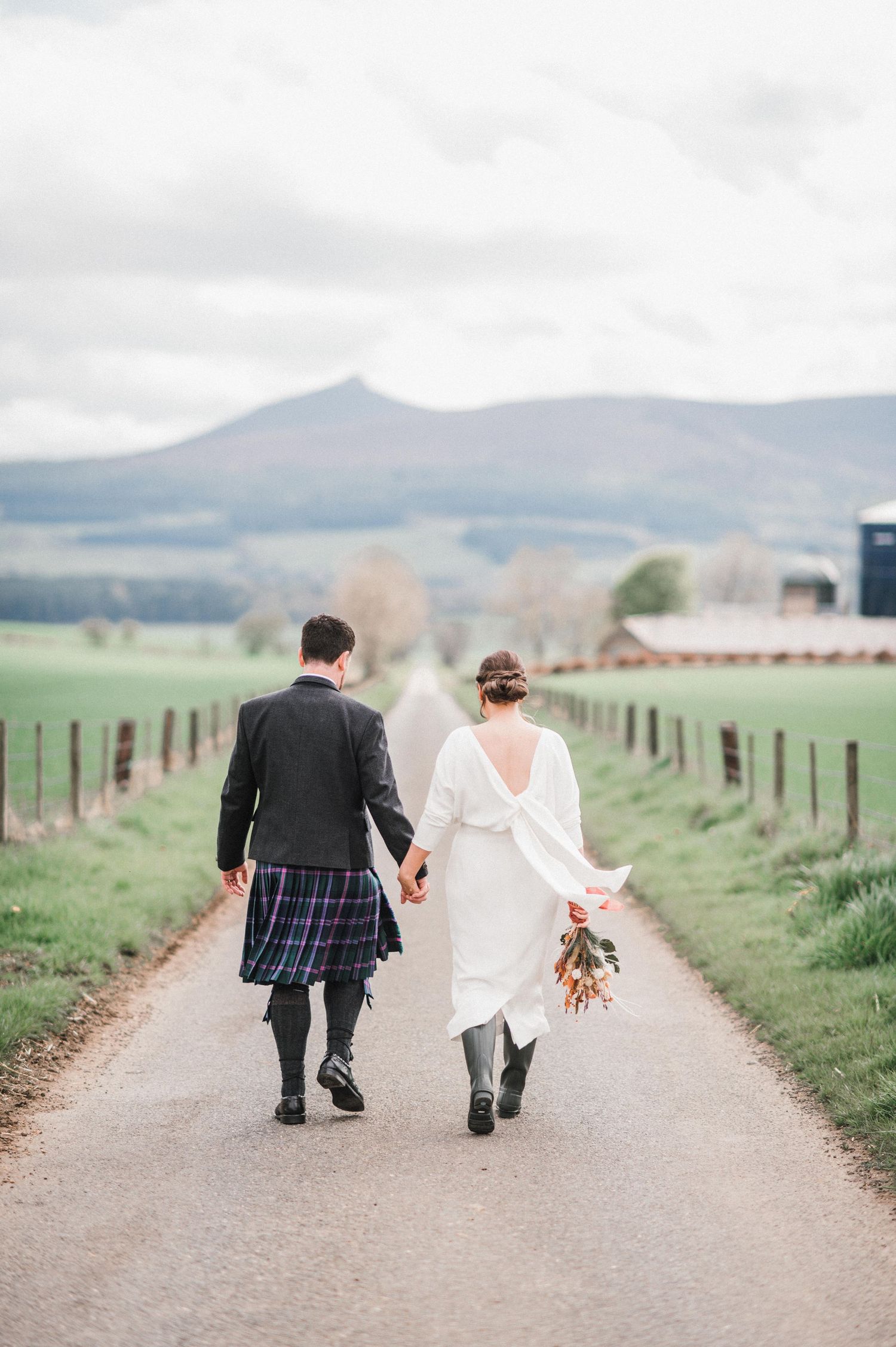 Wedding couple walking down a countryside path with mountain vista and fenced fields.