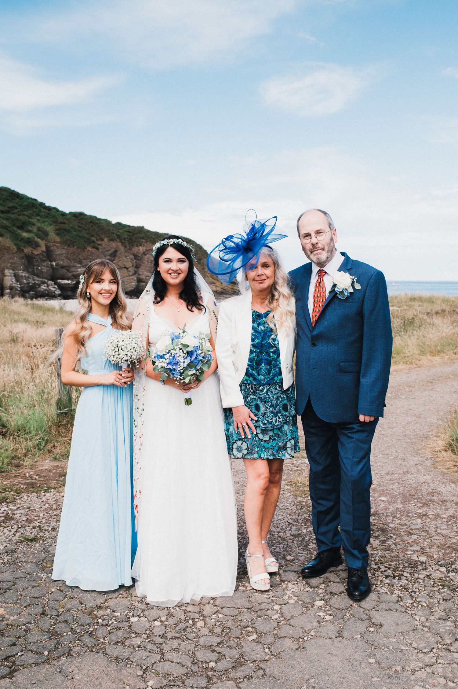 Wedding party in blue and white attire poses on coastal path with cliffs in background.