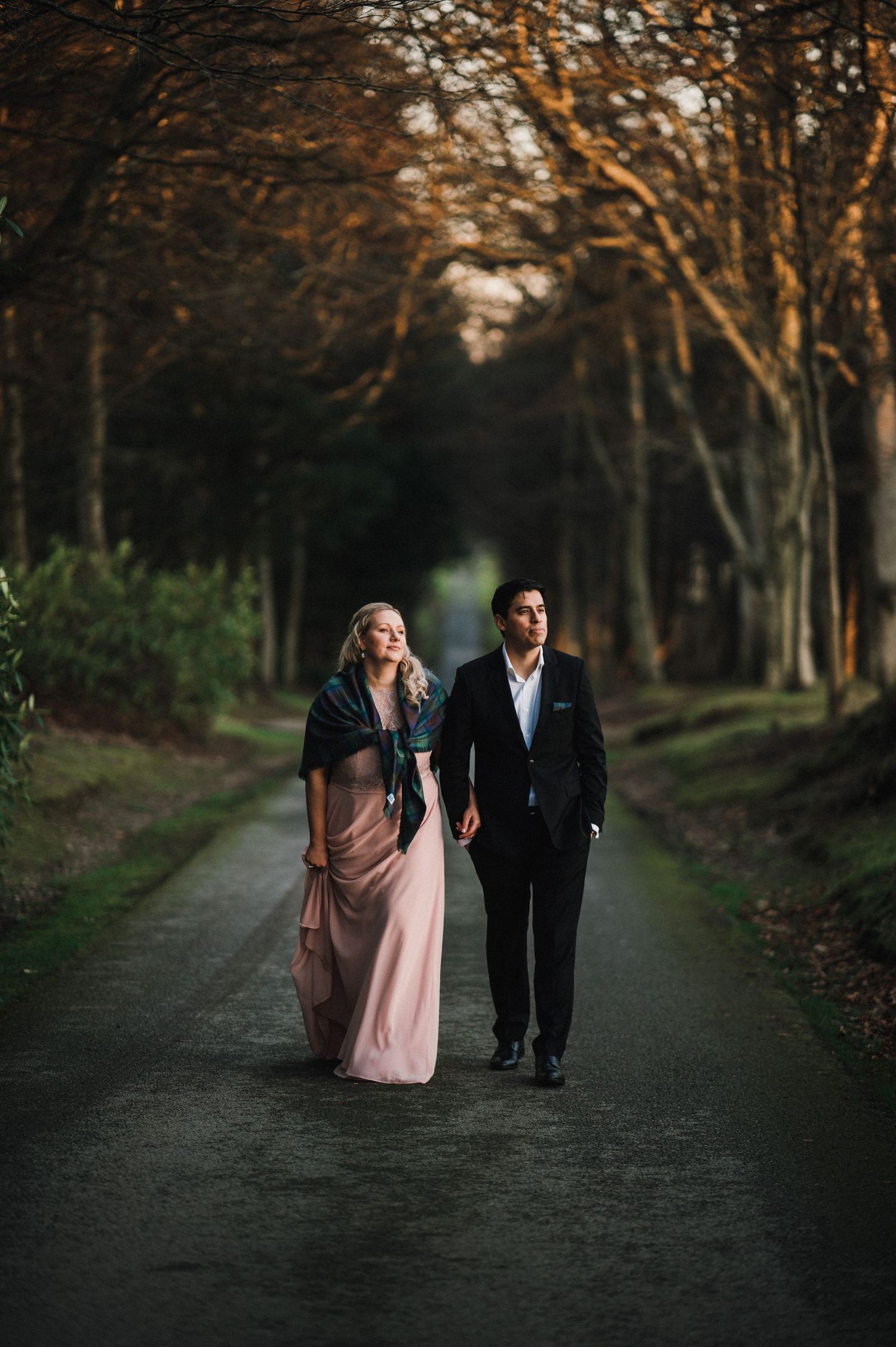 A couple walks along a tree-lined path in formal attire during golden hour.