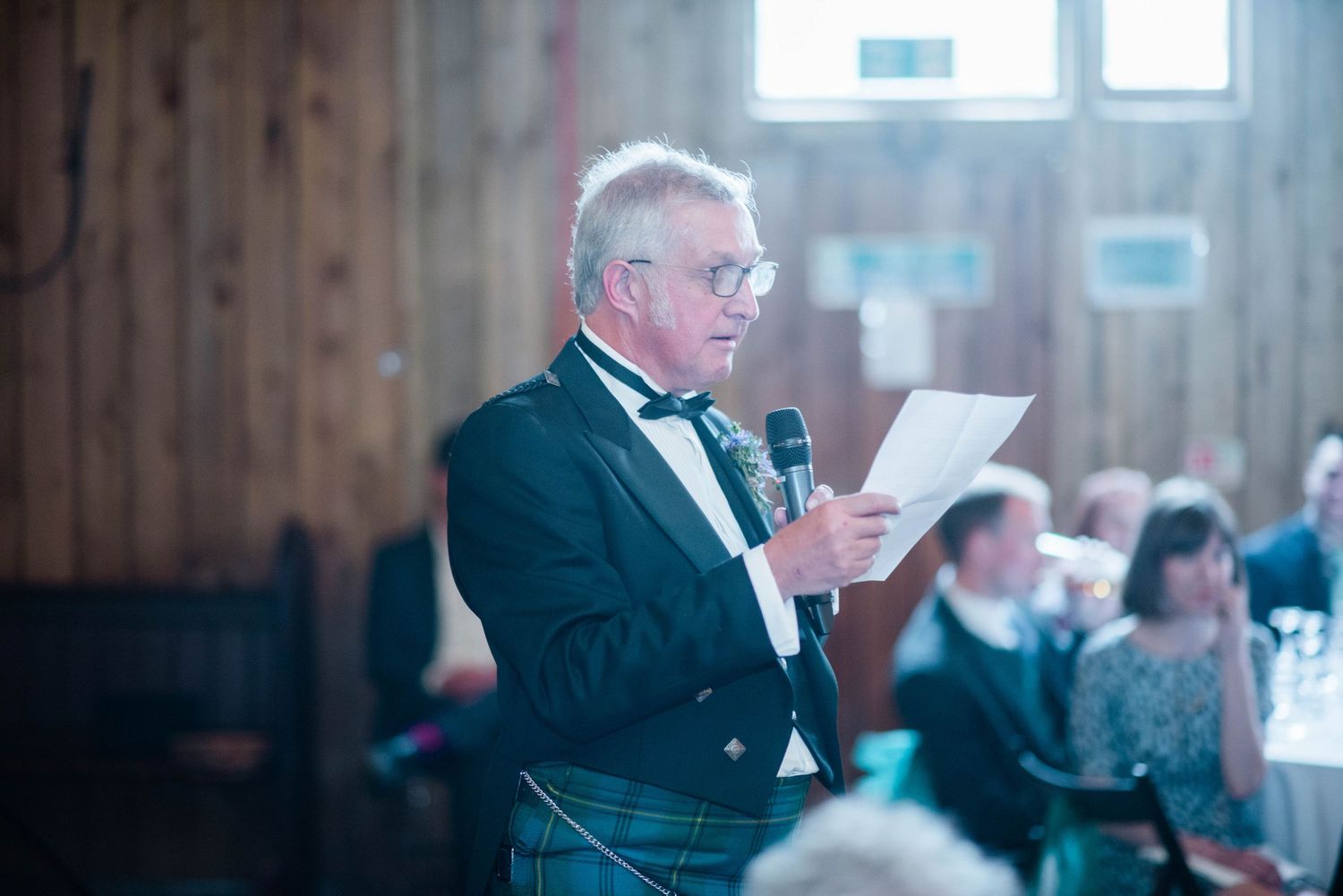 Speaker in formal attire and kilt reads from paper during indoor reception.