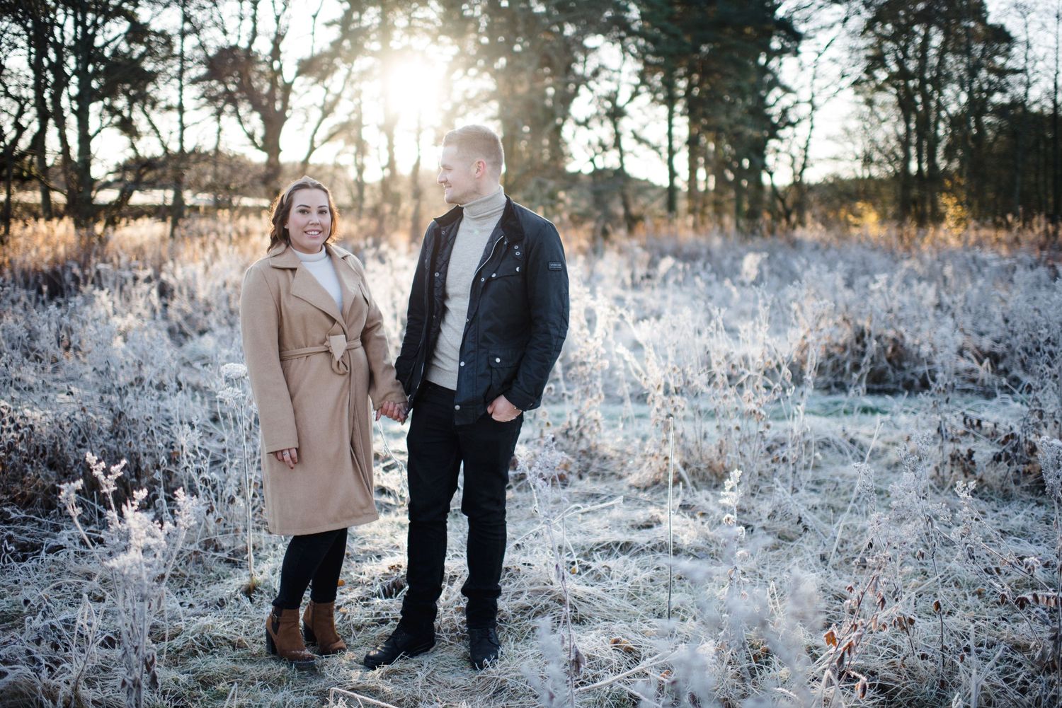 A couple stands together in a frost-covered field during golden hour.