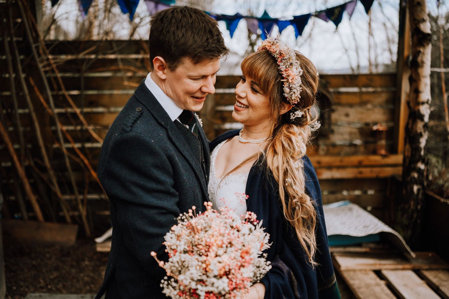 A couple shares an intimate moment at their Scottish wedding, with pink bouquet and traditional highland dress.