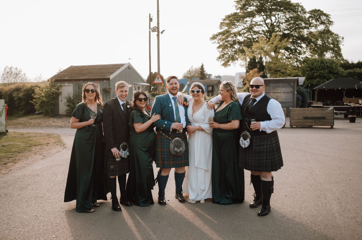 Wedding party poses together outdoors wearing kilts and formal attire in Scottish tradition.