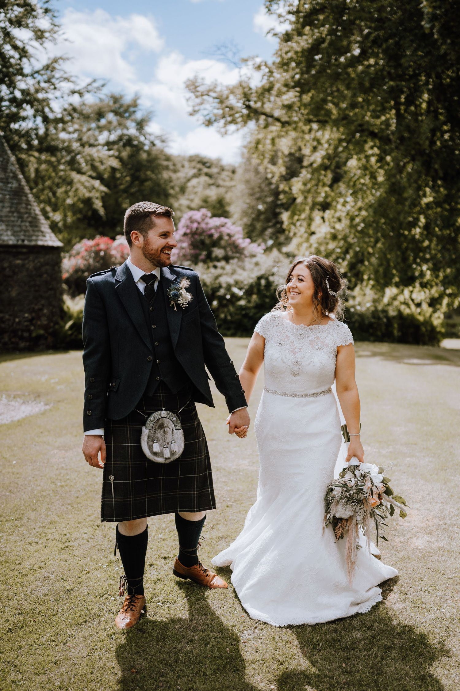 Newlyweds hold hands while walking along garden path in Scottish wedding attire.