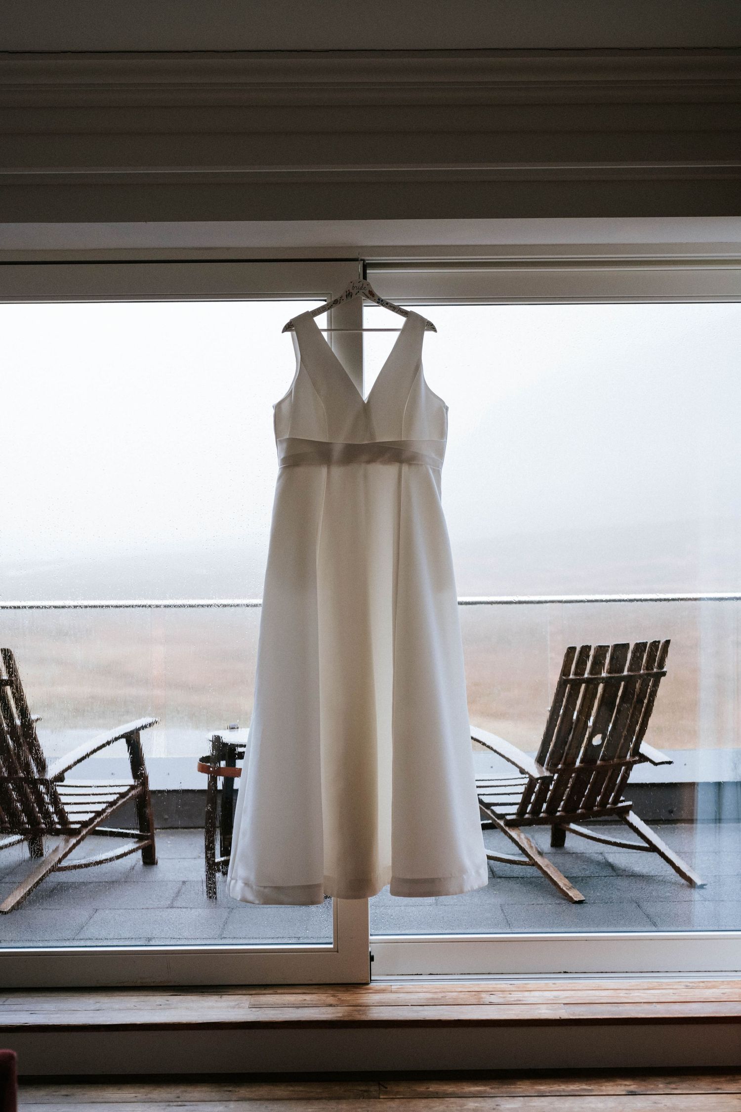 An elegant white wedding dress hanging in front of a window with rocking chairs on a porch.