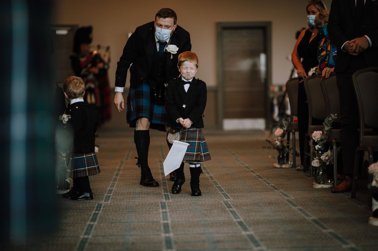 Adult in traditional Scottish kilt walks with young child in matching attire down indoor aisle.