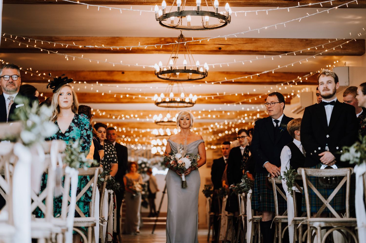 Wedding ceremony processional in rustic venue with twinkling lights and chandeliers overhead.