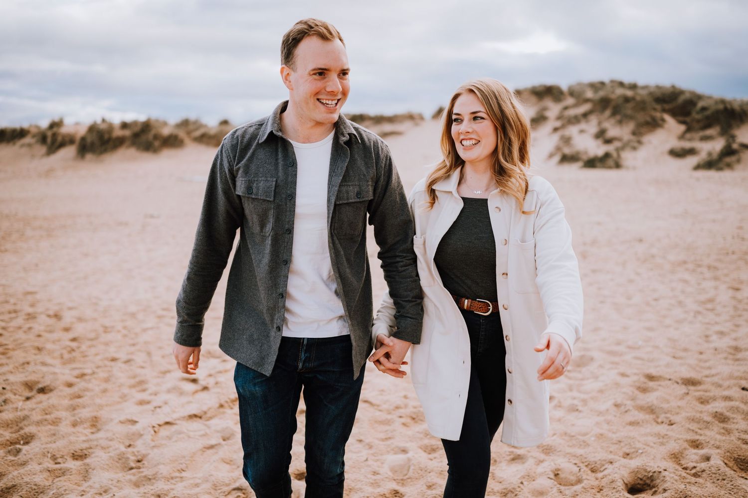 Couple walking hand in hand on sandy beach dunes under cloudy skies.