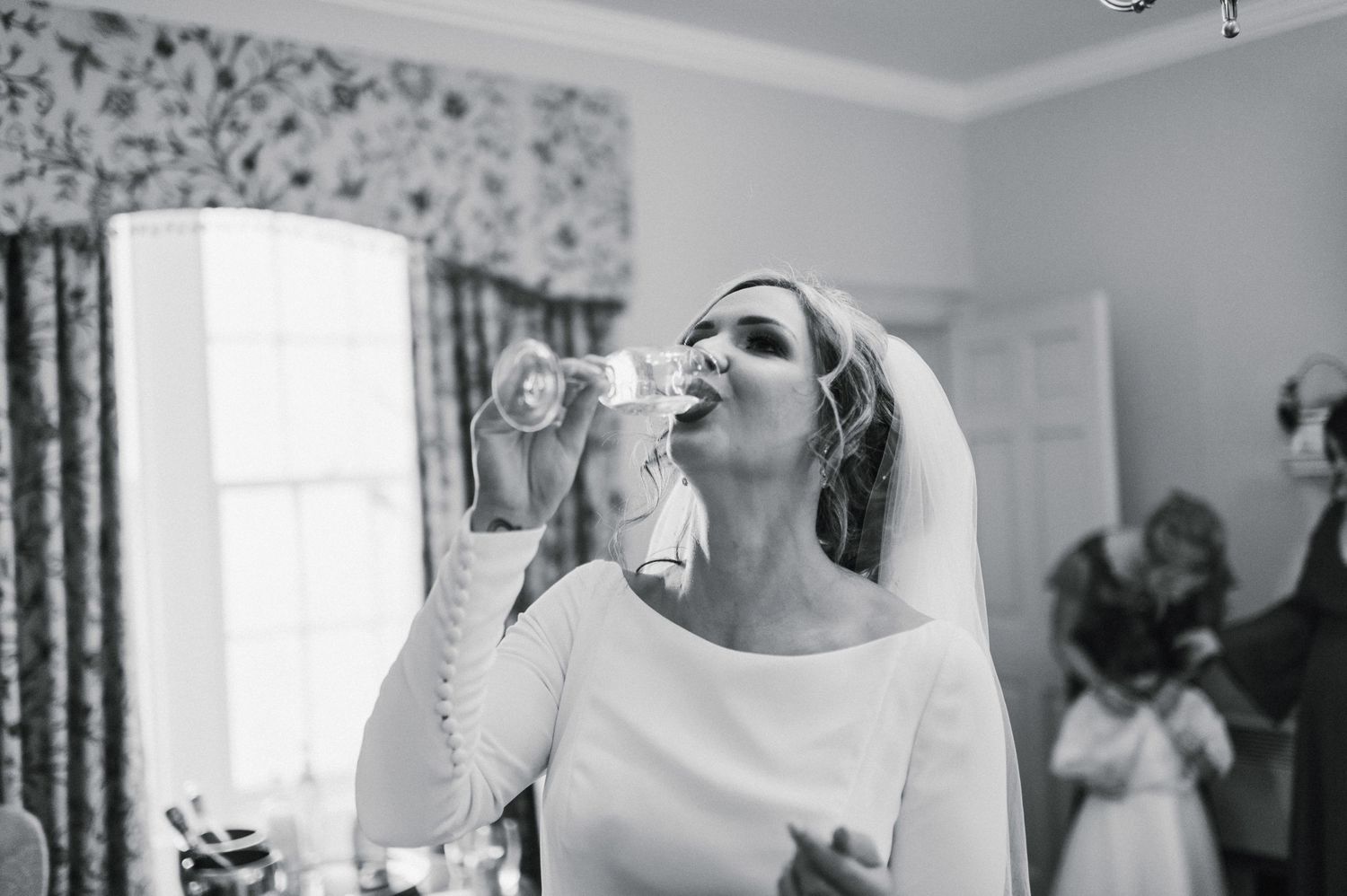 Black and white candid moment of someone taking a drink while wearing a wedding veil.