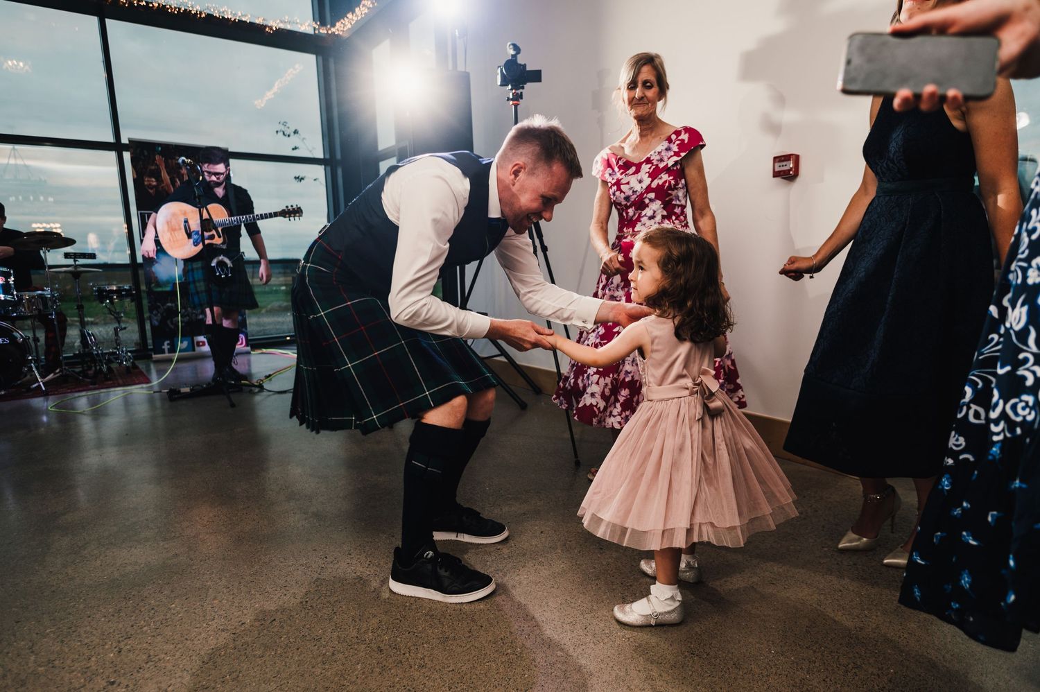 Wedding guest in kilt dances with young guest in pink dress during reception celebrations.