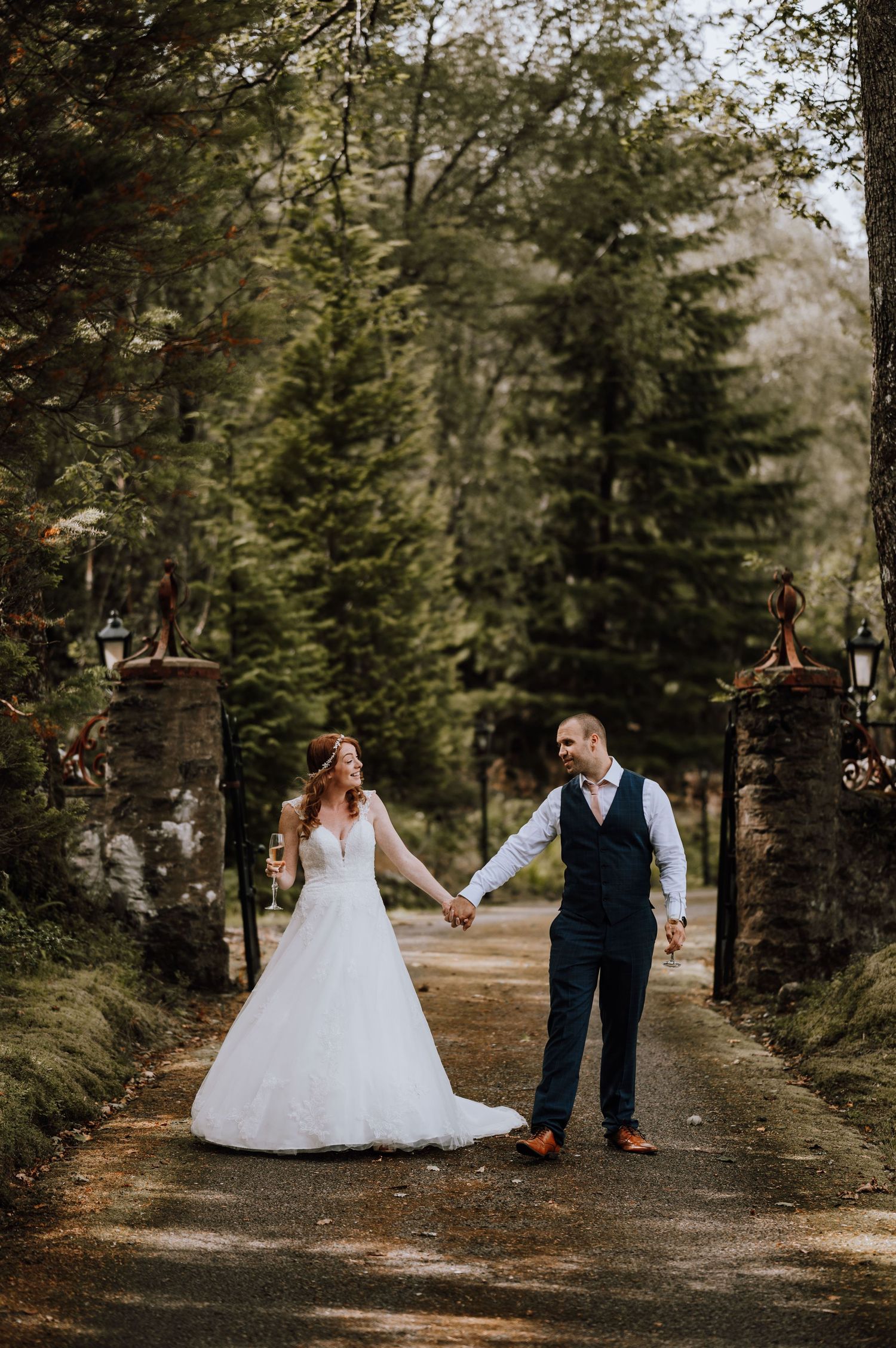 A couple holds hands while walking through a tree-lined stone gate entrance.