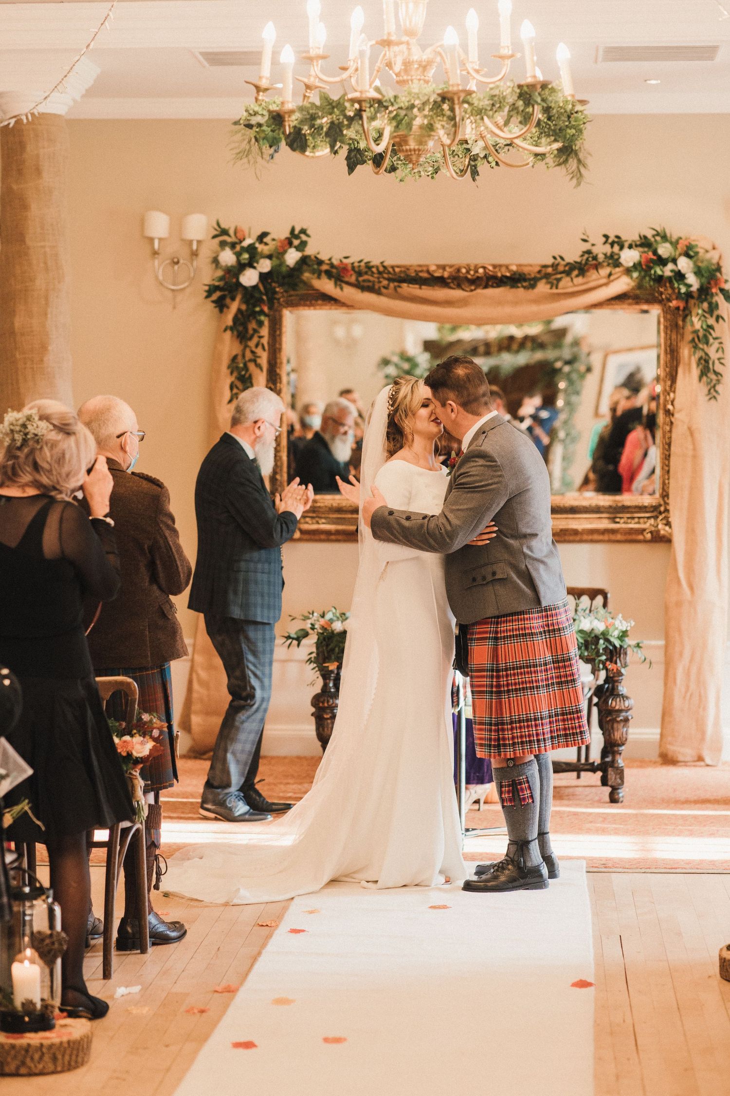 A romantic ceremony moment under a chandelier with floral arch decorations and Scottish Highland attire.