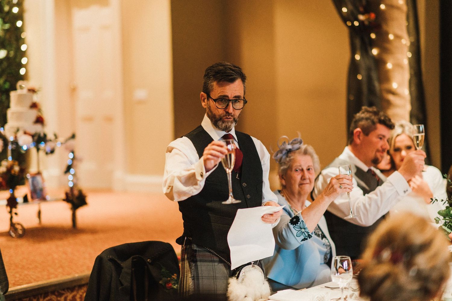 Someone giving a wedding toast during the reception with twinkle lights and warm decor in the background.