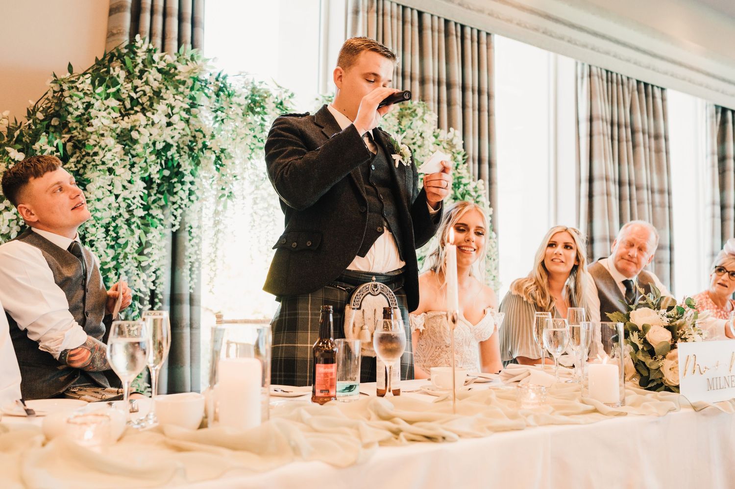 A formal speech being delivered at an elegant wedding reception with greenery decor in the background.