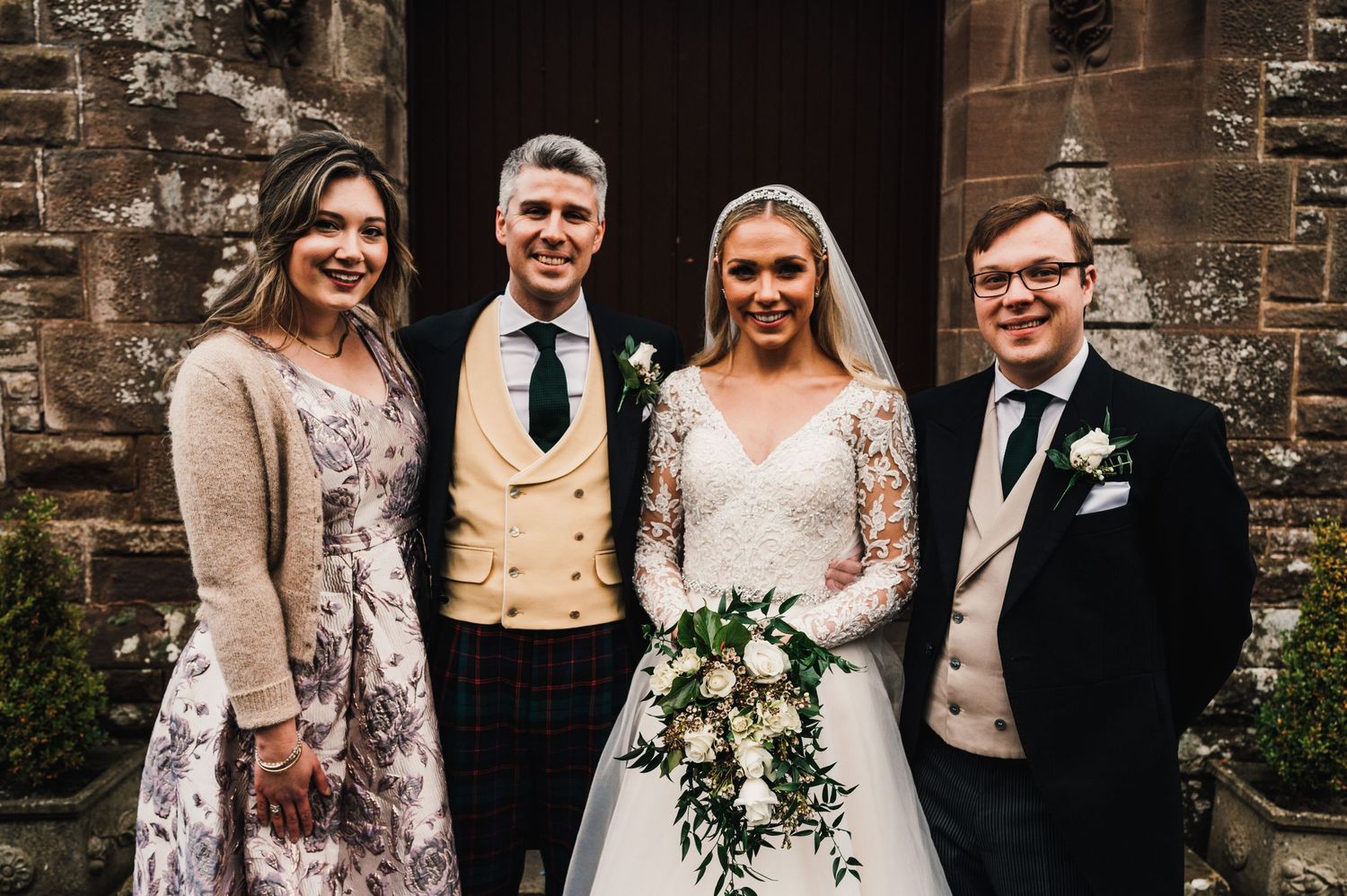 Small wedding party group poses together in formal attire outside a historic church entrance.