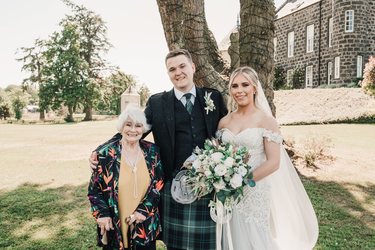 A wedding photo with three people posing outside a stone building.