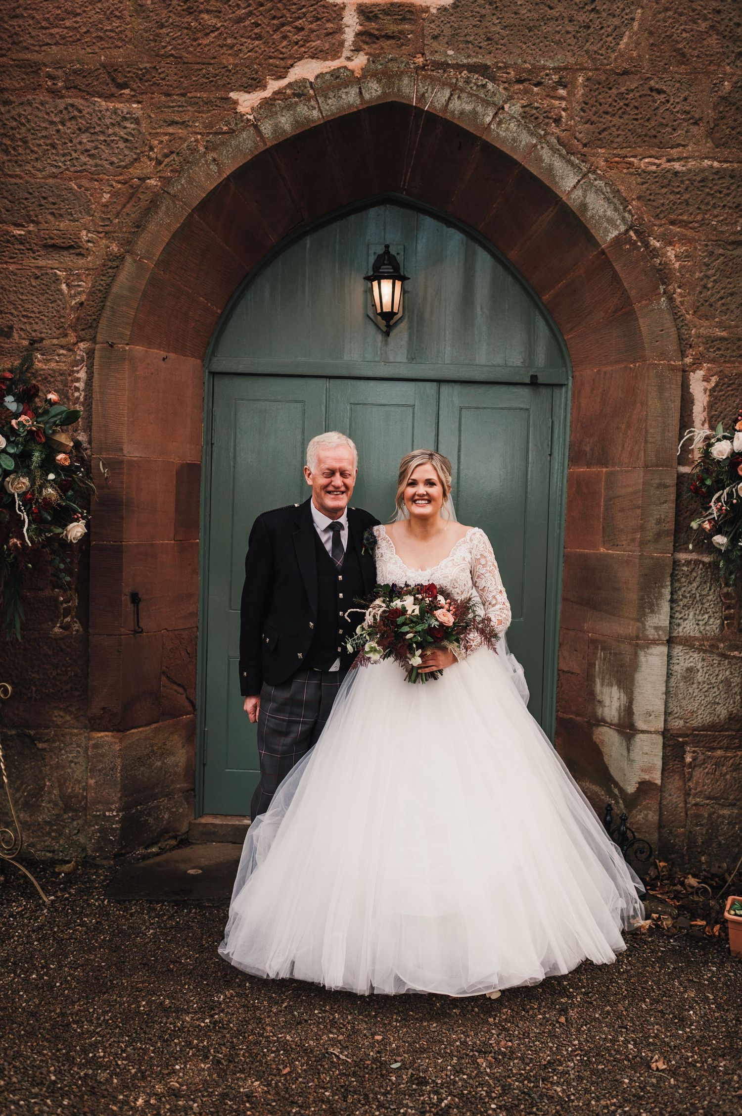 A wedding couple poses in front of a green arched doorway with stone walls and a lantern.