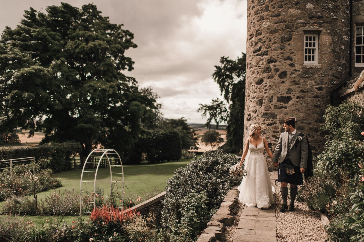 Wedding couple stroll through castle gardens with beautiful landscaping and stone architecture.